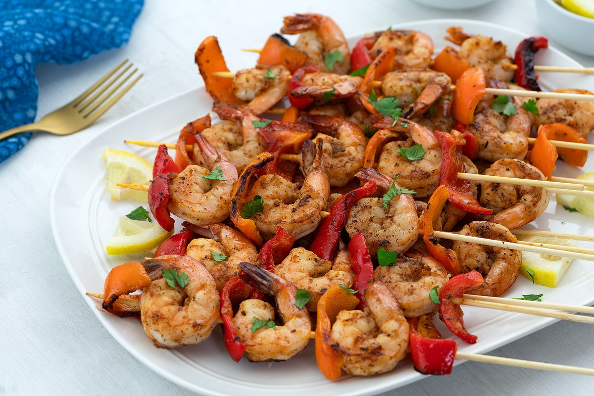 Juicy shrimp skewers on a plate, set on a white table with a blue towel, a golden fork, and a cup of lemon slices nearby.
