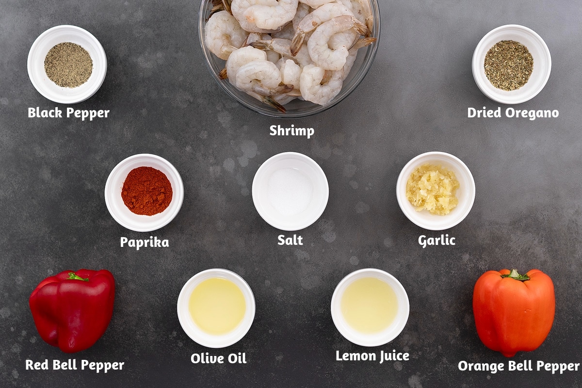 Ingredients for shrimp skewers arranged on a gray table, including black pepper, shrimp, dried oregano, paprika, salt, garlic, red and orange bell peppers, olive oil, and lemon juice.