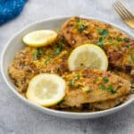 Lemon pepper chicken breasts in a white bowl on a white table, with a blue towel, and a golden fork around it.