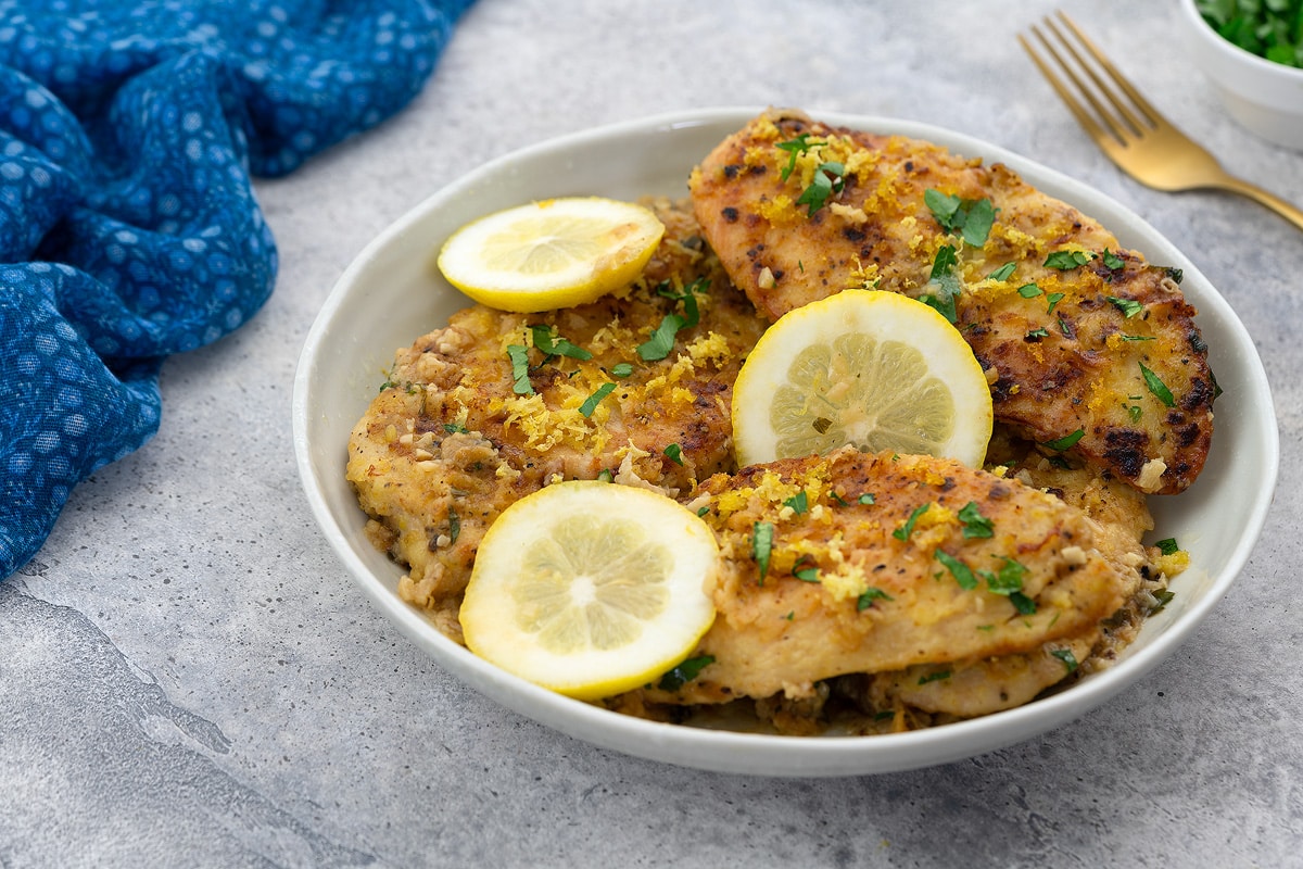 Lemon pepper chicken breasts in a white bowl on a white table, with a blue towel, a golden fork, and a cup of parsley around it.