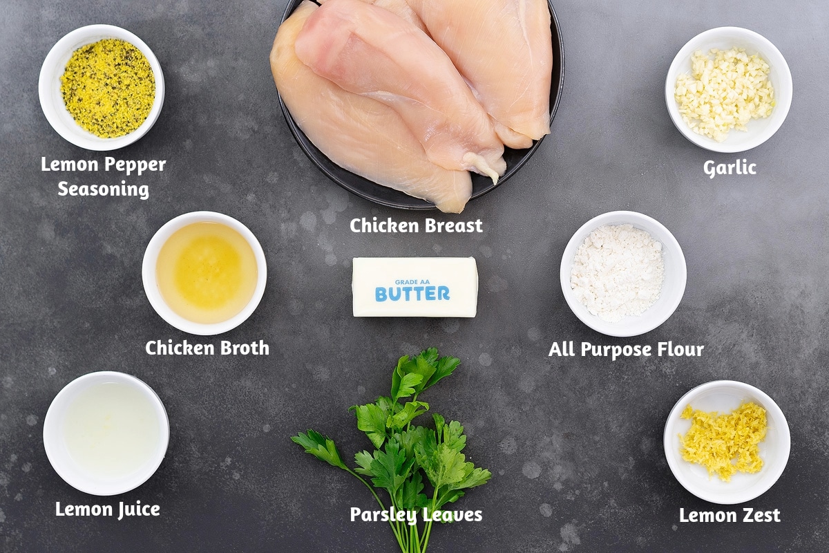 Ingredients for lemon pepper chicken on a gray table, including lemon pepper seasoning, chicken breast, garlic, chicken broth, butter, all-purpose flour, lemon juice, parsley leaves, and lemon zest.