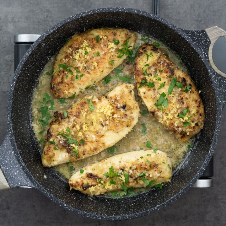 Lemon pepper chicken in savory sauce garnished with lemon zest and parsley leaves in a pan.