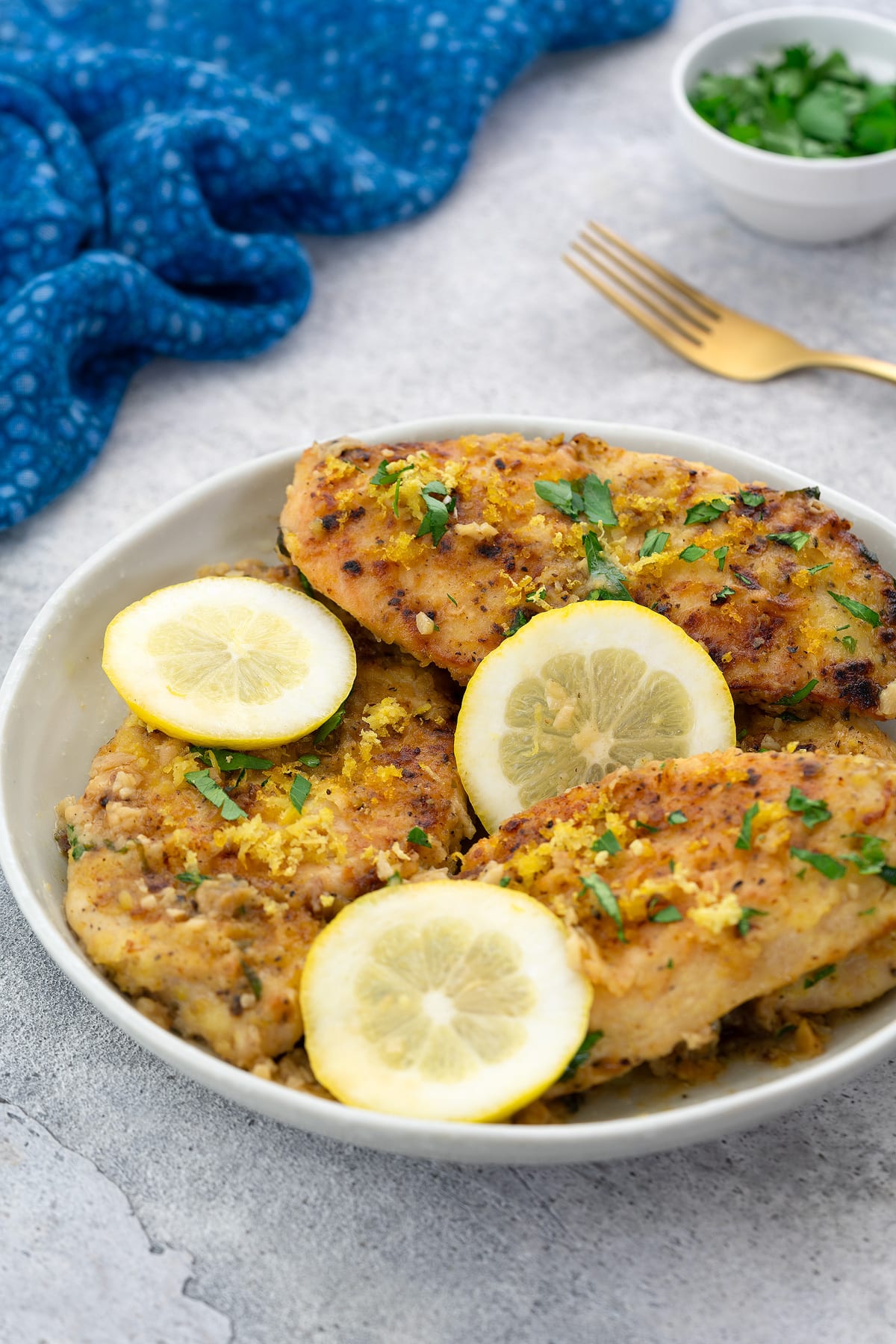 Lemon pepper chicken breasts in a white bowl on a white table, with a blue towel, a golden fork, and a cup of parsley around it.