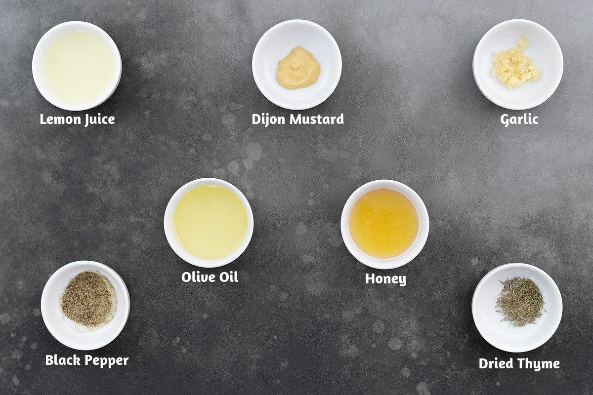 Lemon Vinaigrette ingredients arranged on a gray table, including lemon juice, Dijon mustard, garlic, olive oil, honey, black pepper powder, and dried thyme.