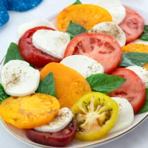A colorful Caprese salad in an oval plate on a white table, accompanied by a blue towel, a golden fork, and a small cup of ground black pepper.