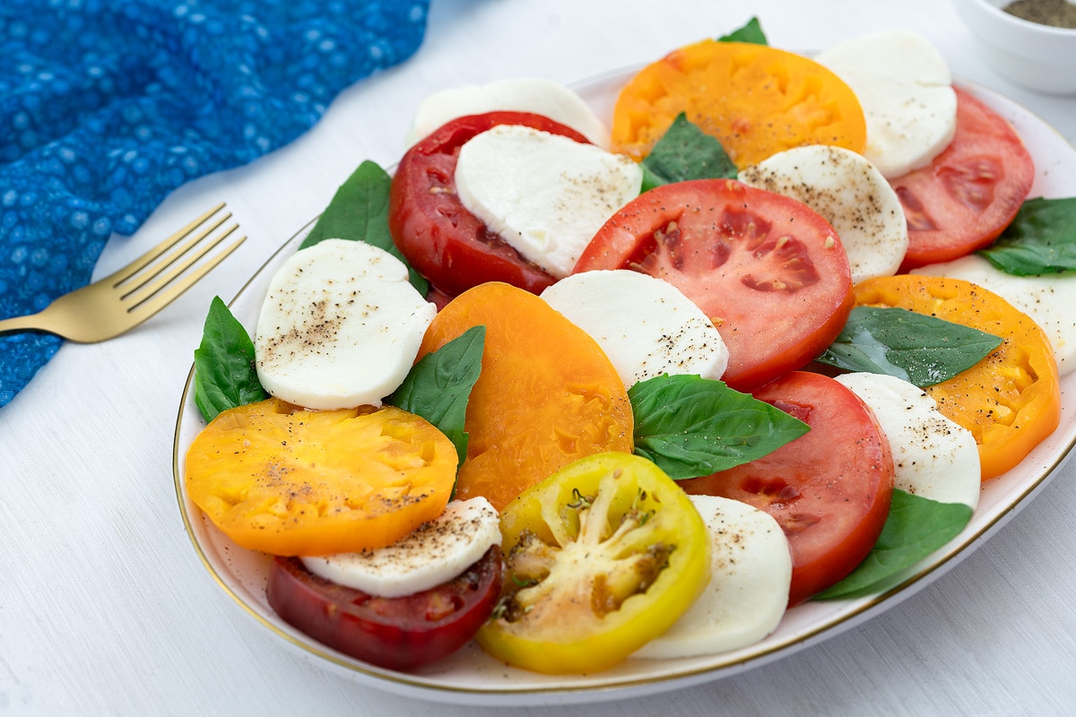 A colorful Caprese salad in an oval plate on a white table, accompanied by a blue towel, a golden fork, and a small cup of ground black pepper.