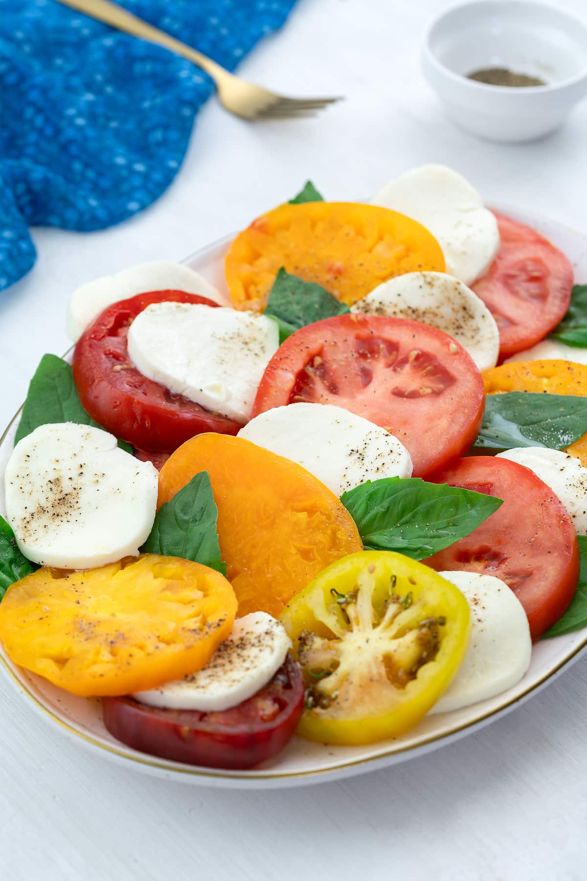 A colorful Caprese salad in an oval plate on a white table, accompanied by a blue towel, a golden fork, and a small cup of ground black pepper.