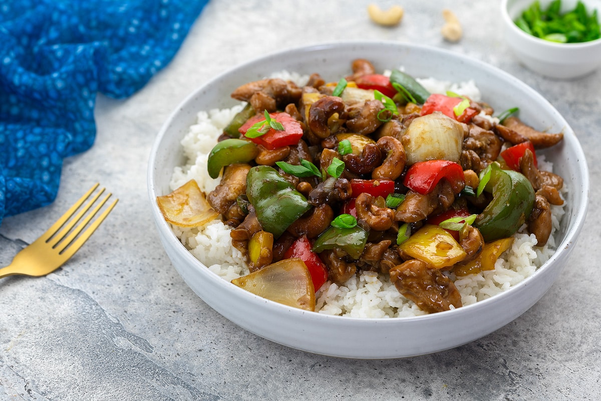 Chinese cashew chicken served in a white bowl with rice on a white table, surrounded by a blue towel, golden fork, and a cup of green onions.