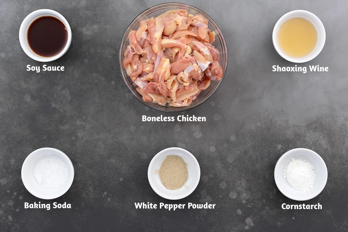 Ingredients for cashew chicken placed on a gray table: soy sauce, boneless chicken, Shaoxing wine, baking soda, white pepper powder, and cornstarch.