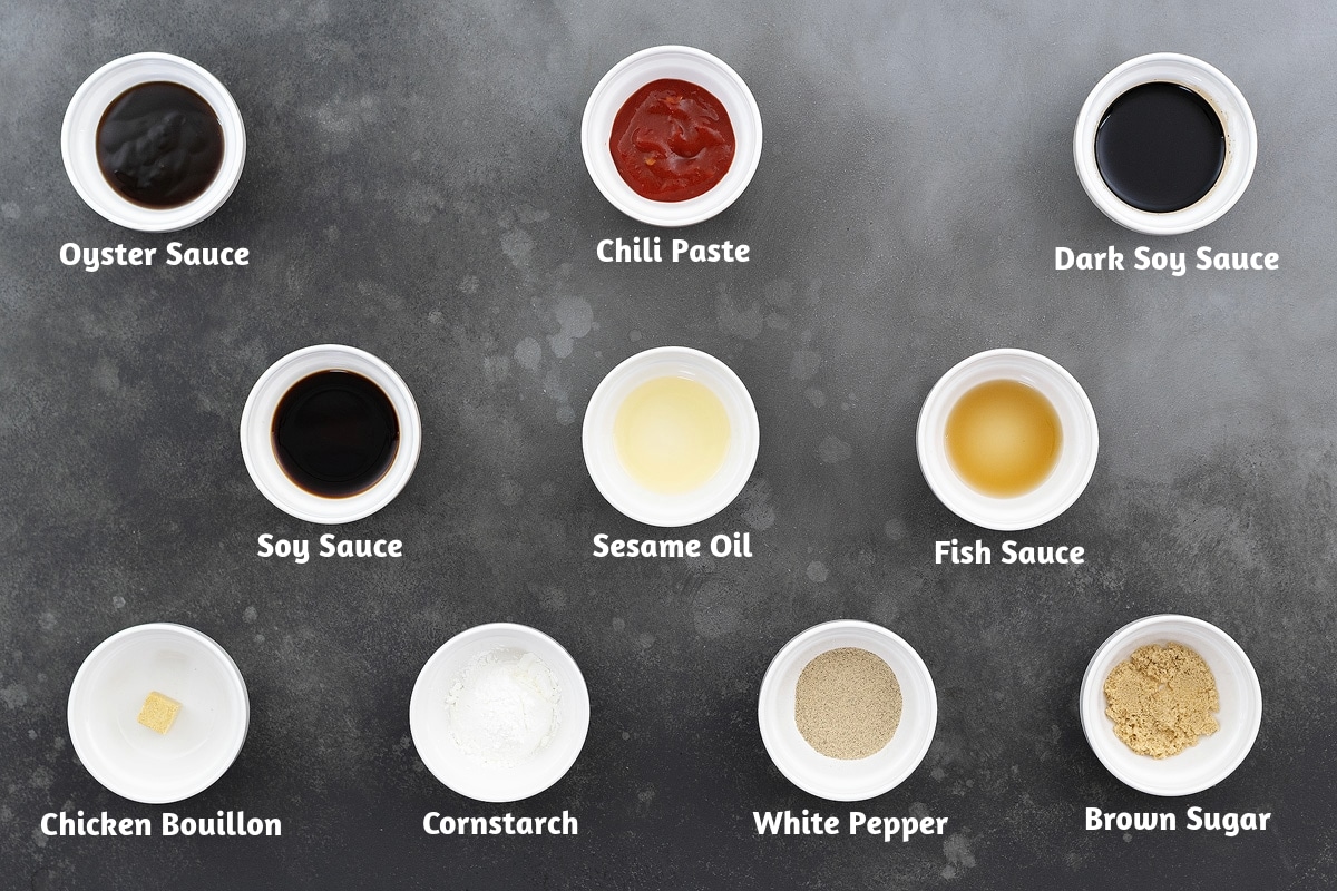 Ingredients for cashew chicken sauce arranged on a gray table: oyster sauce, chili paste, dark soy sauce, soy sauce, sesame oil, fish sauce, chicken bouillon, cornstarch, white pepper powder, and brown sugar.