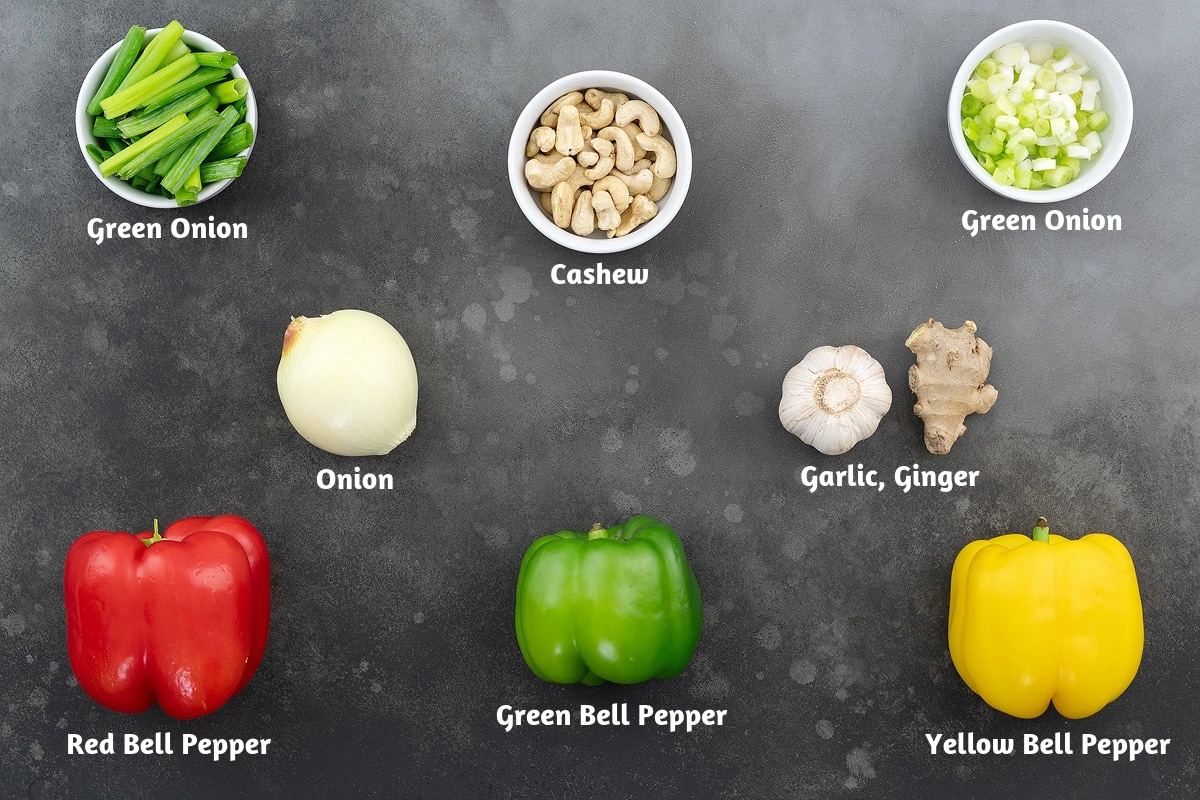 Ingredients for stir frying cashew chicken on a gray table: green onion (green part), cashews, green onion (white part), onion, garlic, ginger, red bell pepper, green bell pepper, and yellow bell pepper.