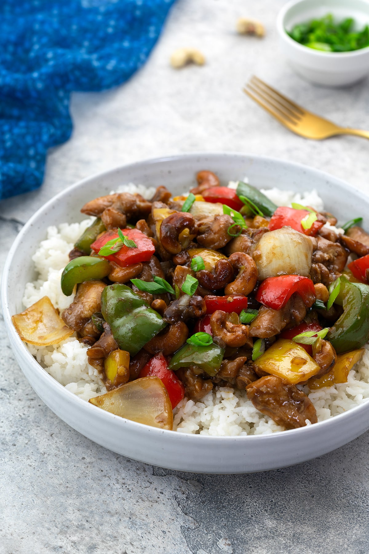 Chinese cashew chicken served in a white bowl with rice on a white table, surrounded by a blue towel, golden fork, and a cup of green onions.