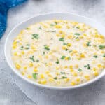 Homemade creamed corn in a white bowl on a white table with a blue towel nearby.