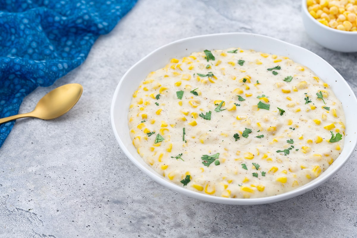 Homemade creamed corn in a white bowl on a white table, with a blue towel, golden spoon, and a small cup of corn kernels nearby.