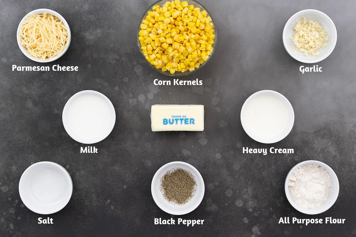 Creamed corn ingredients on a gray table: Parmesan cheese, corn kernels, garlic, milk, butter, heavy cream, salt, black pepper powder, and all-purpose flour.