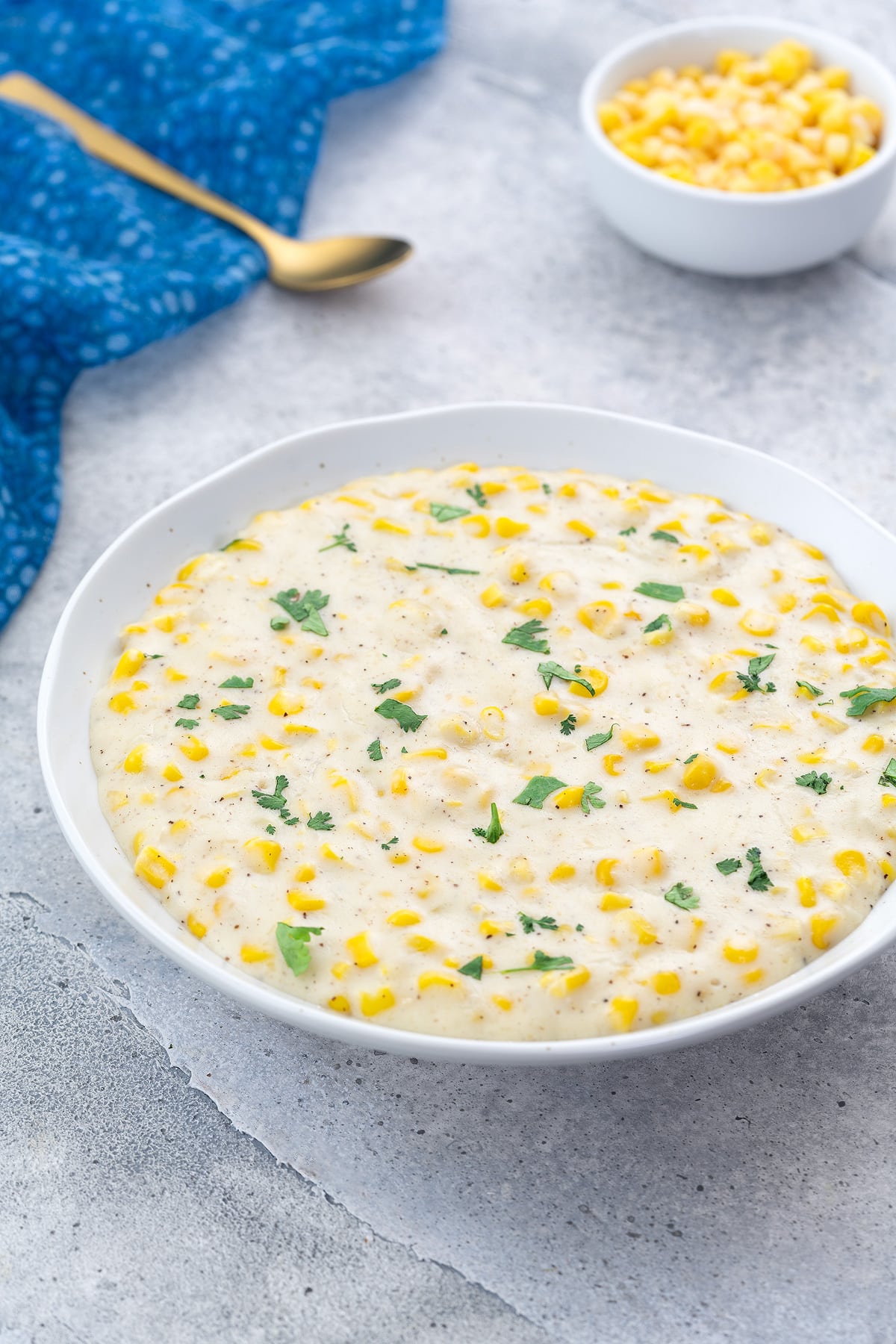 Homemade creamed corn in a white bowl on a white table, with a blue towel, golden spoon, and a small cup of corn kernels nearby.