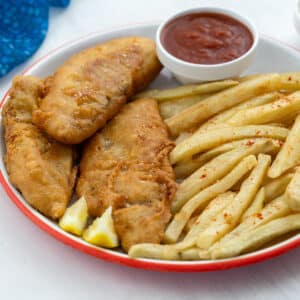 A plate of homemade fish and chips with ketchup on a white table. A blue towel and a small bowl of mayo are nearby.