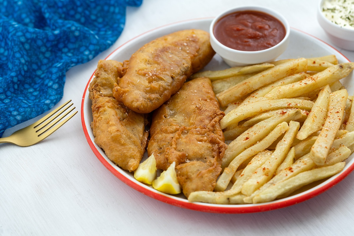 A plate of homemade fish and chips with ketchup, on a white table. A blue towel, fork, and a small bowl of mayo are nearby.