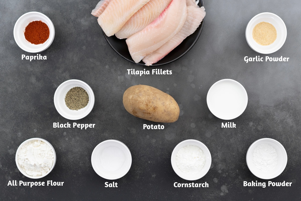 Ingredients for fish and chips on a gray table, including paprika, tilapia fillets, garlic powder, black pepper, potato, milk, all-purpose flour, salt, cornstarch, and baking powder.