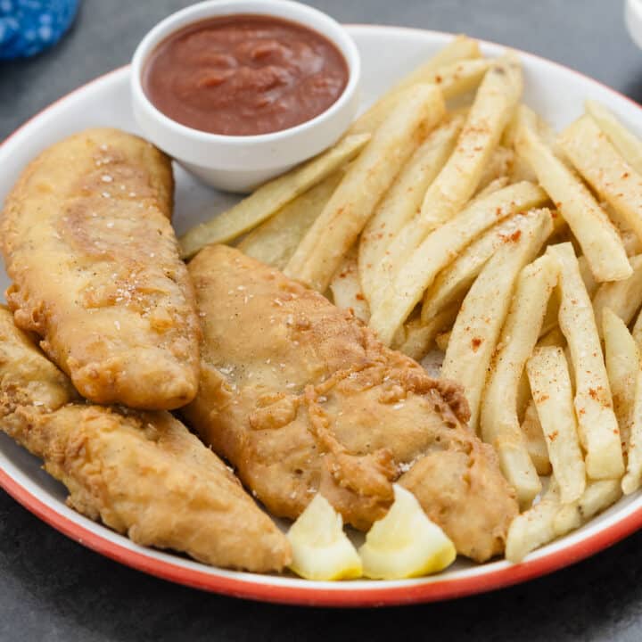 Fish and chips served on a white plate along with lemon wedges and ketchup.