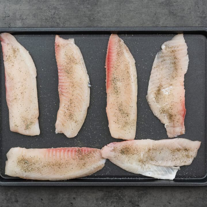 A large tray with tilapia fillets seasoned with black pepper and salt.