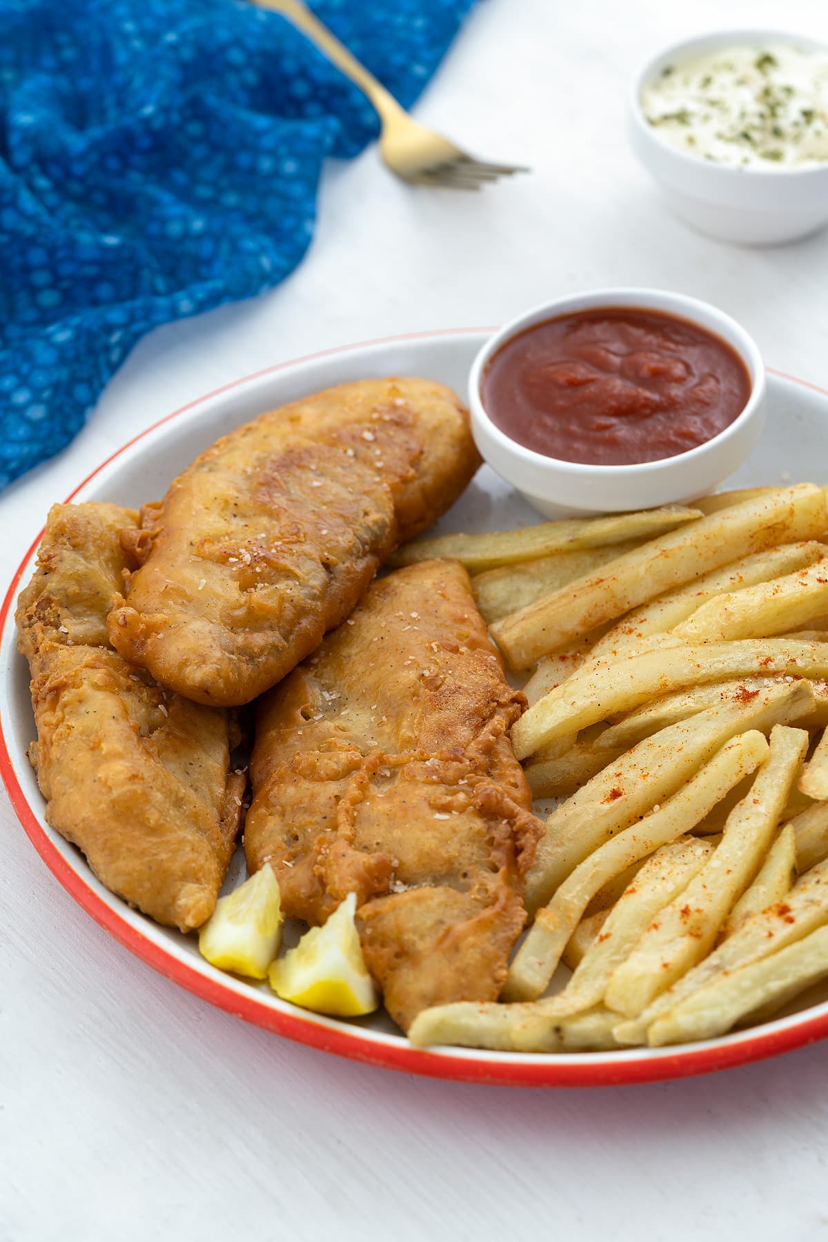 A plate of homemade fish and chips with ketchup, on a white table. A blue towel, fork, and a small bowl of mayo are nearby.