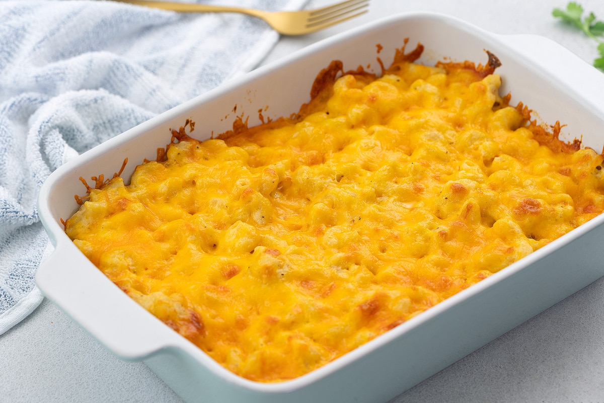 Baked mac and cheese in a white baking dish on a white table, with a white towel, golden fork, and mint leaves around it.