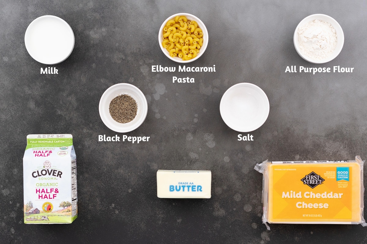 Ingredients for mac and cheese on a gray table: milk, elbow macaroni pasta, all-purpose flour, black pepper powder, salt, half and half, butter, and cheddar cheese.