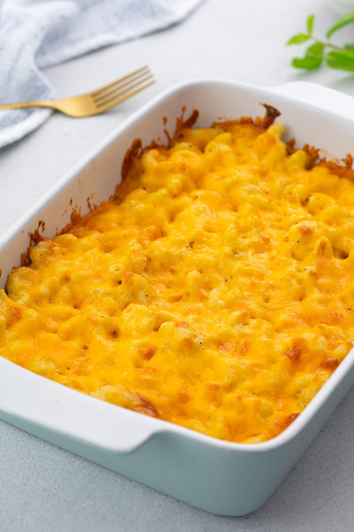 Baked mac and cheese in a white baking dish on a white table, with a white towel, golden fork, and mint leaves around it.