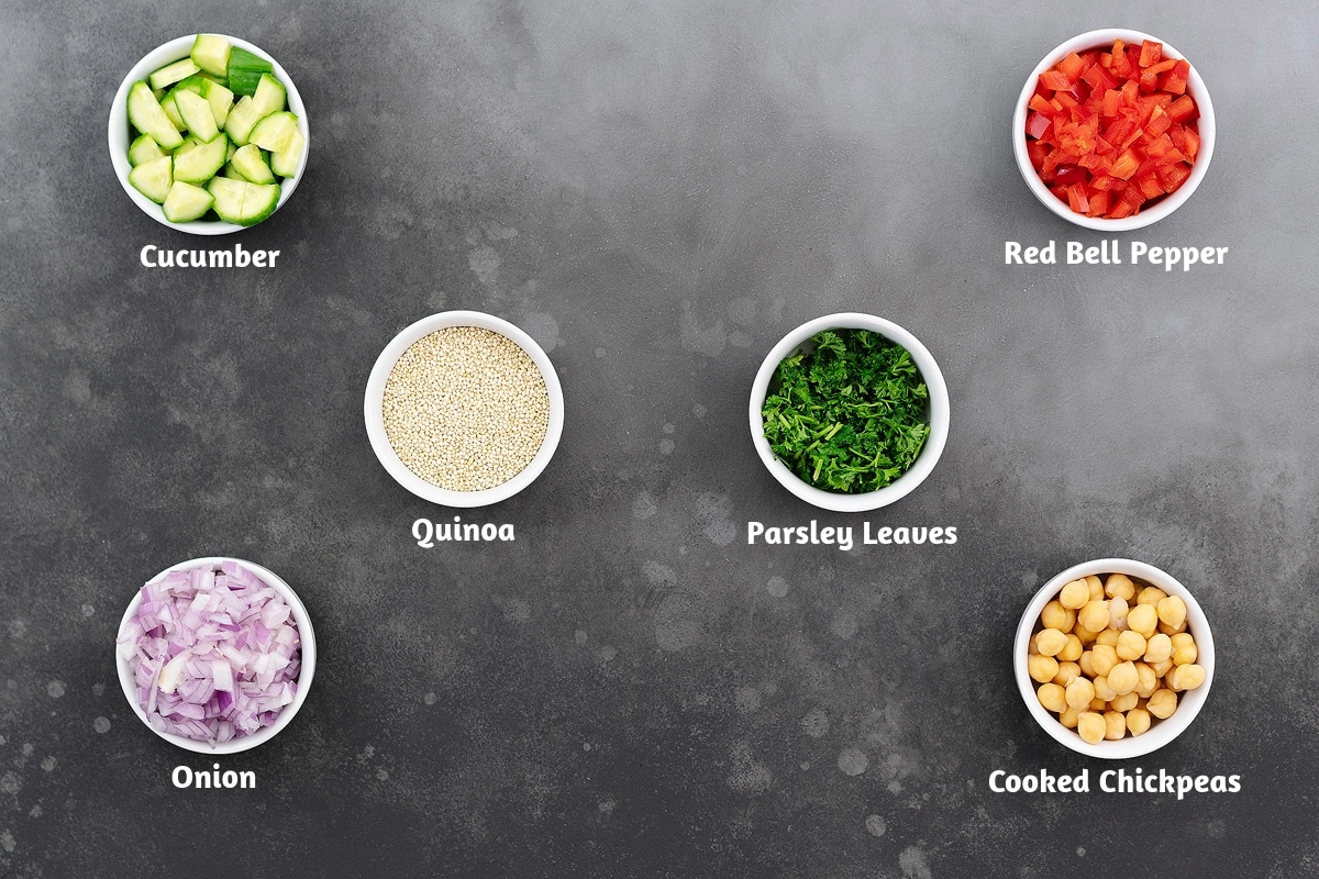 Ingredients for quinoa salad neatly arranged on a gray table: slices of cucumber, chopped red bell pepper, cooked quinoa, fresh parsley leaves, chopped onion, and cooked chickpeas.