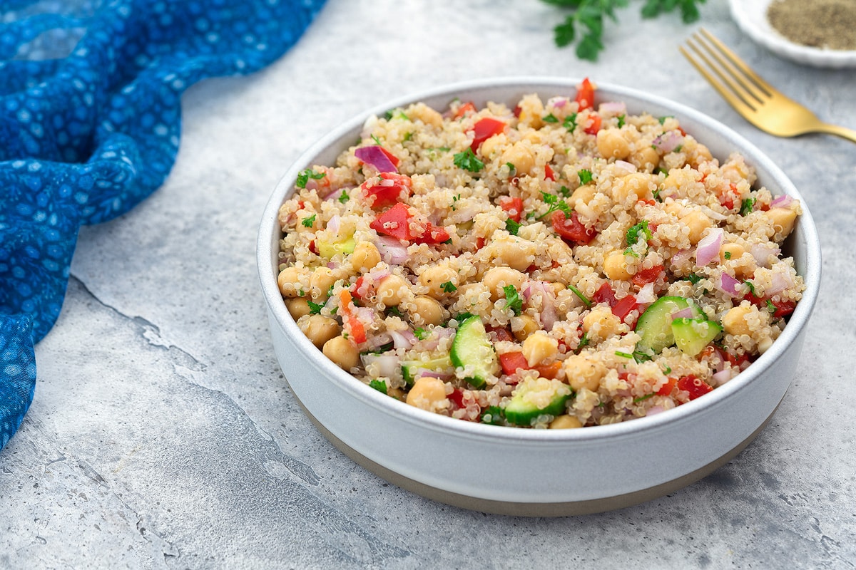 Colorful quinoa salad served in a white bowl on a white table, accompanied by a blue towel, a golden fork, a small plate of black pepper powder, and some fresh cilantro scattered around.