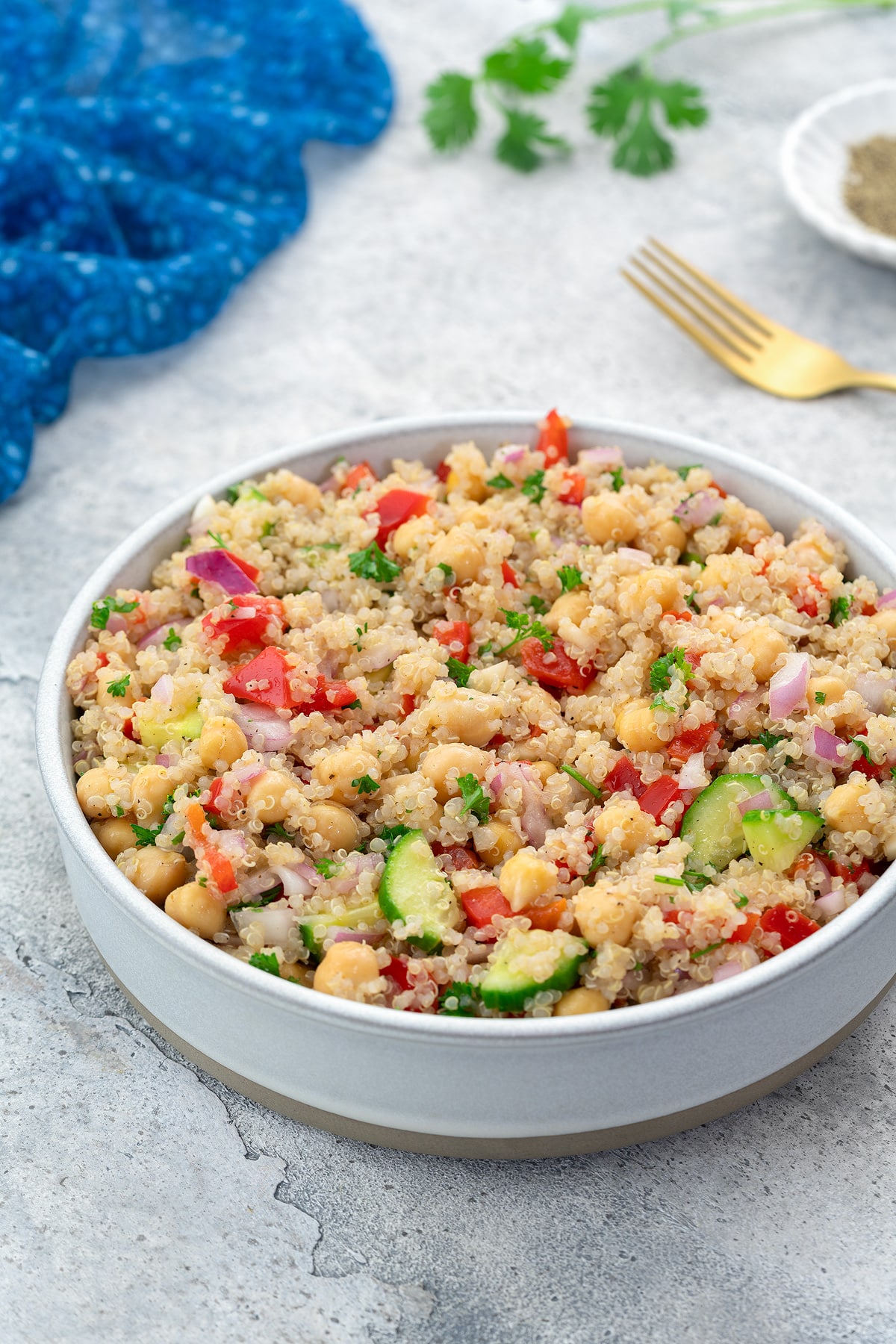 Colorful quinoa salad served in a white bowl on a white table, accompanied by a blue towel, a golden fork, a small plate of black pepper powder, and some fresh cilantro scattered around.