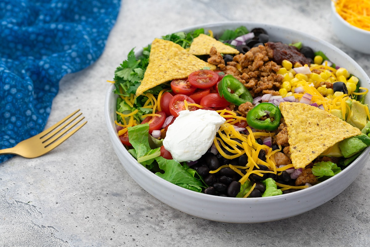 Homemade taco salad in a white bowl on a white table with a blue towel, golden fork, and a cup of cheddar cheese nearby.