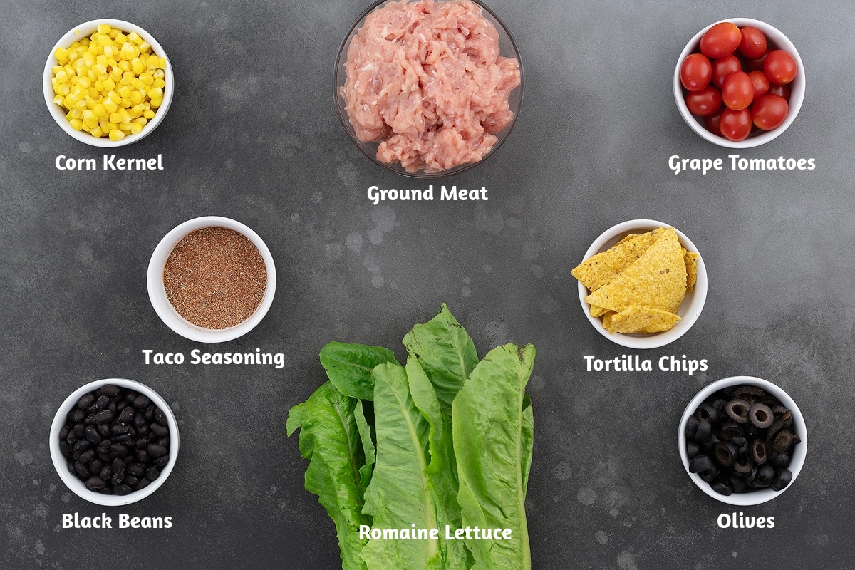 Taco salad ingredients on a gray table: corn kernels, ground meat, grape tomatoes, taco seasoning, tortilla chips, black beans, romaine lettuce, and olives.