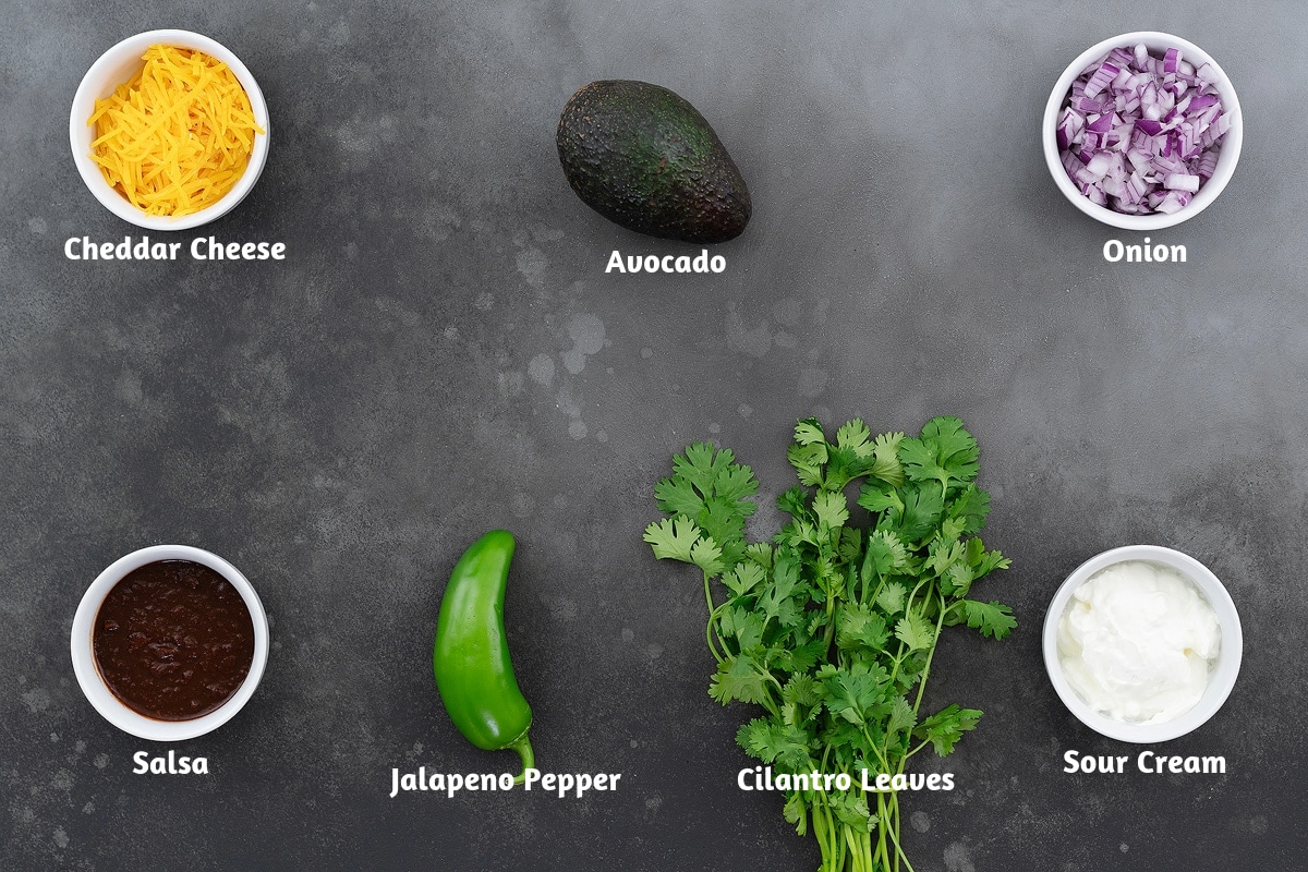 Taco salad ingredients on a gray table: cheddar cheese, avocado, onion, salsa, jalapeno pepper, cilantro leaves, and sour cream.