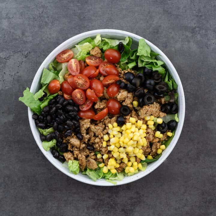 A bowl with lettuce, ground meat, tomatoes, beans, olives, and corn layered.