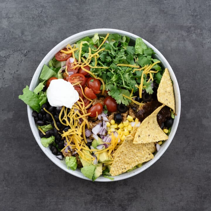A bowl with taco salad ingredients layered and topped with cheese, cilantro, sour cream, salsa, and tortilla chips.