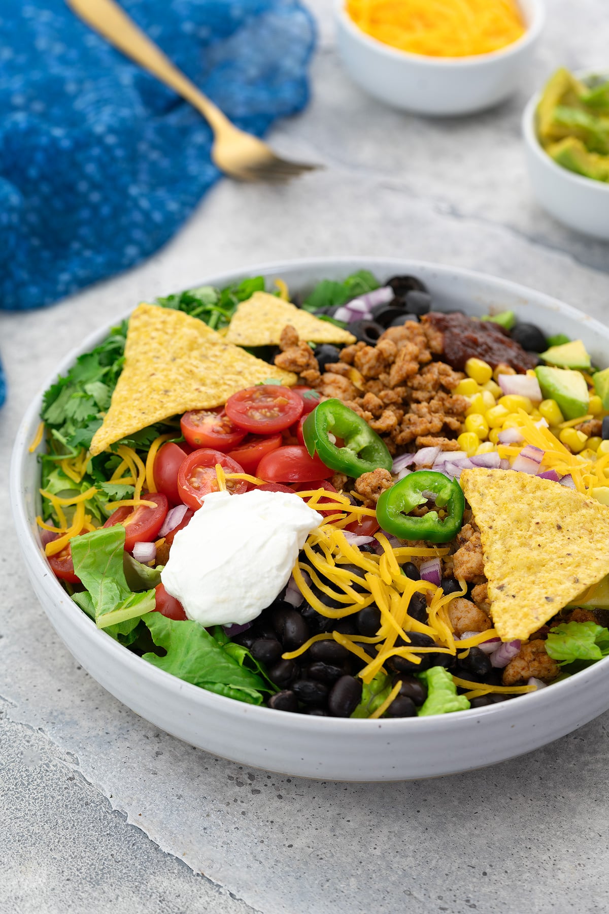 Homemade taco salad in a white bowl on a white table with a blue towel, golden fork, cup of cheddar cheese, and avocado nearby.