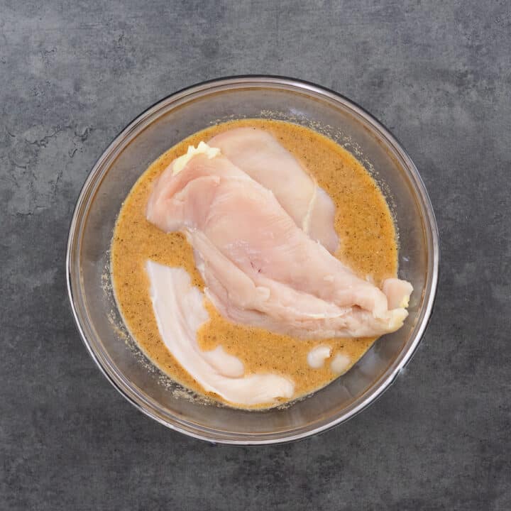 A bowl with marinade and boneless chicken breast pieces.