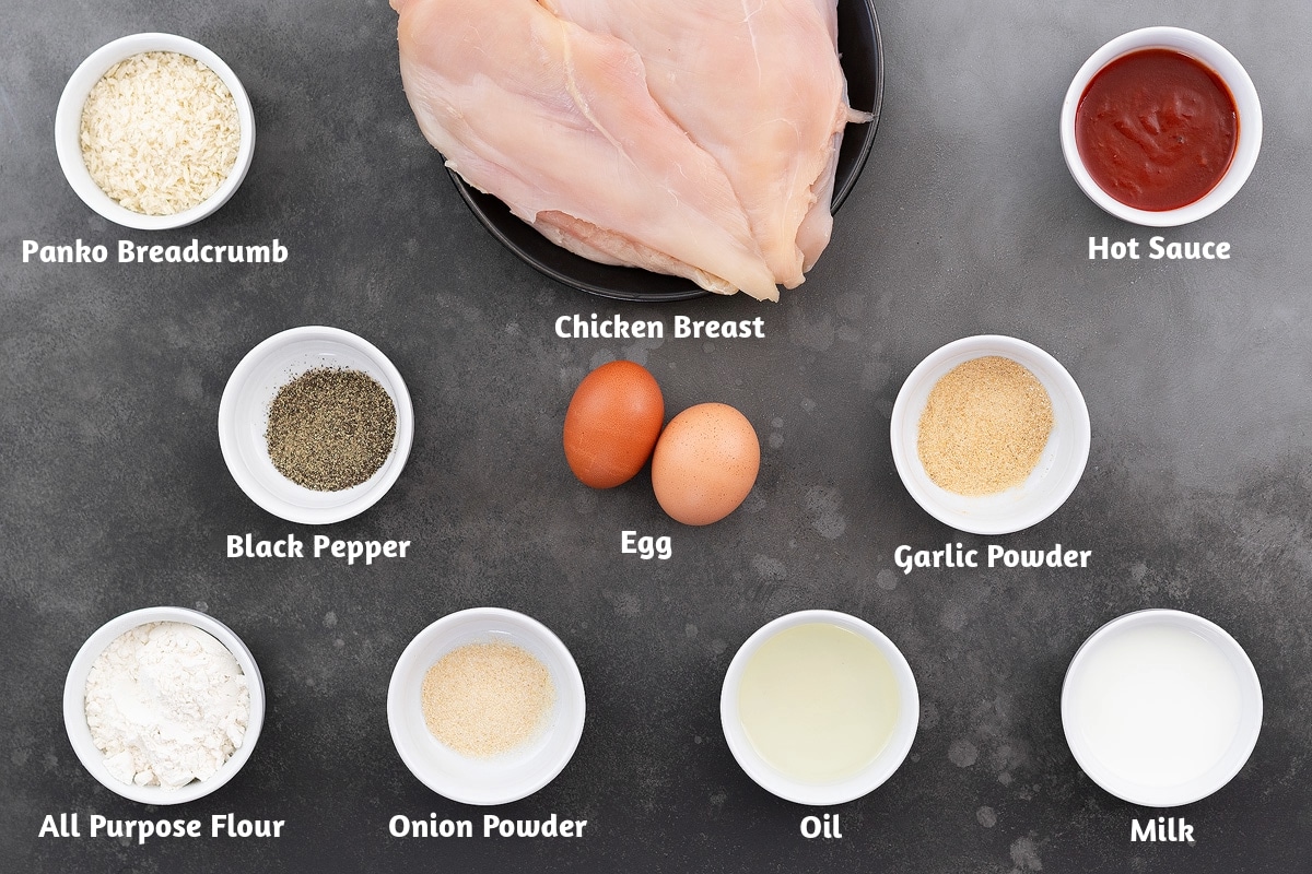 Chicken fried chicken recipe ingredients on a gray table: panko breadcrumbs, chicken breast, hot sauce, black pepper powder, egg, garlic powder, all-purpose flour, onion powder, oil, and milk.