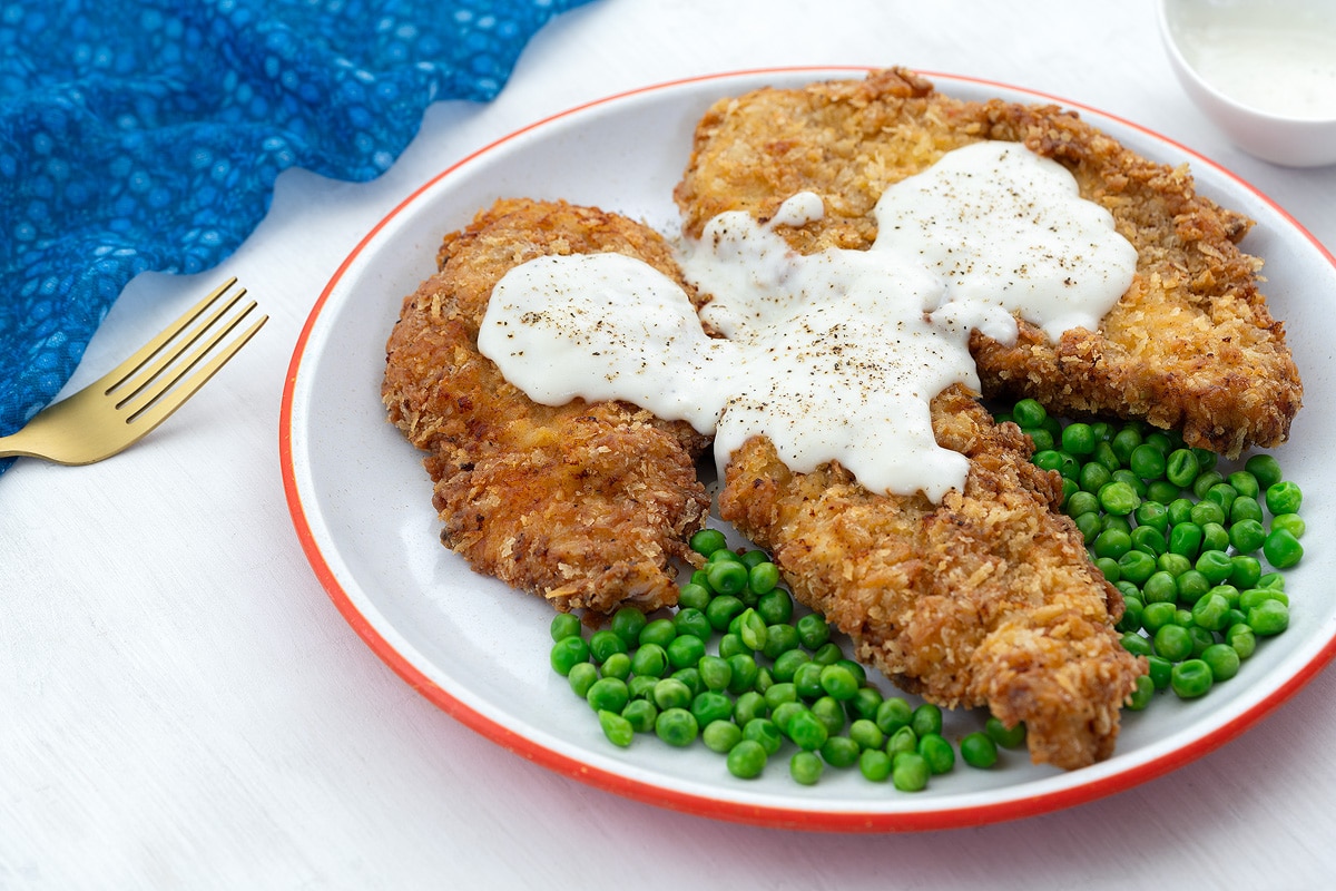 Homemade chicken fried chicken breasts and green peas on a white plate with a blue towel, golden fork, and a cup of gravy nearby on a white table.