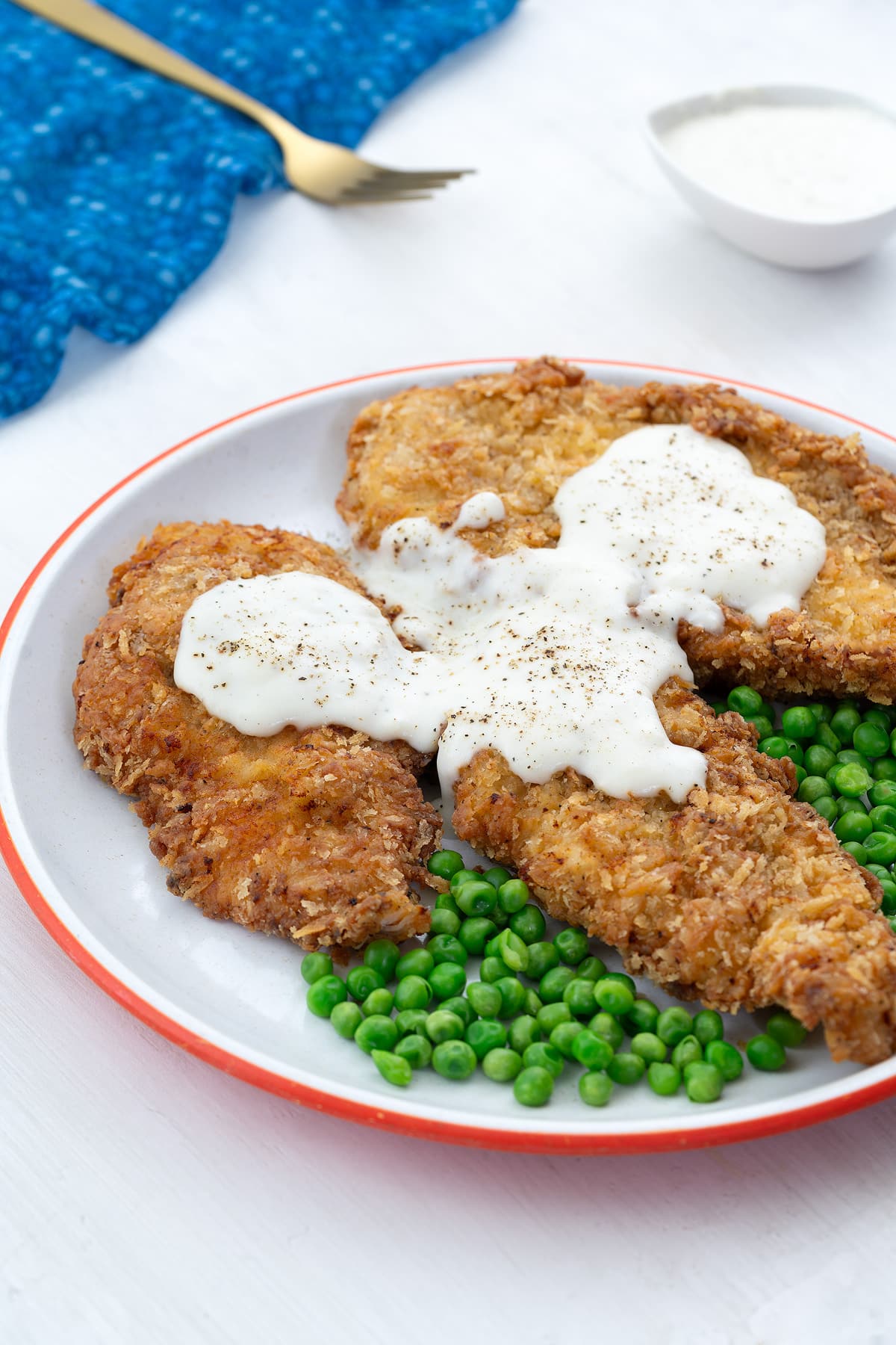 Homemade chicken fried chicken breasts and green peas on a white plate with a blue towel, golden fork, and a cup of gravy nearby on a white table.