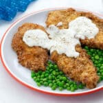 Homemade chicken fried chicken breasts and green peas on a white plate with a blue towel nearby on a white table.