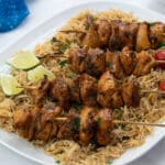 Homemade chicken skewers on a white plate with rice, a blue towel on a white table.