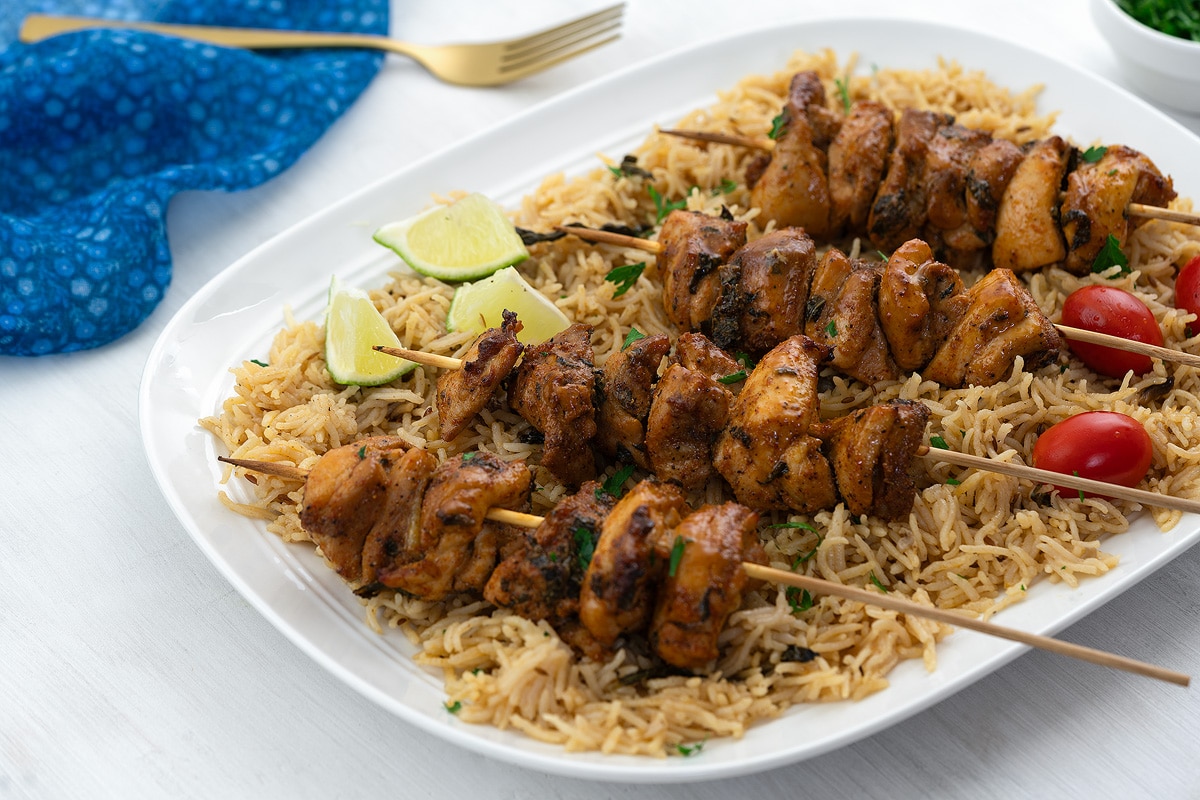 Homemade chicken skewers on a white plate with rice, a golden fork, a blue towel, and a small cup of cilantro on a white table.