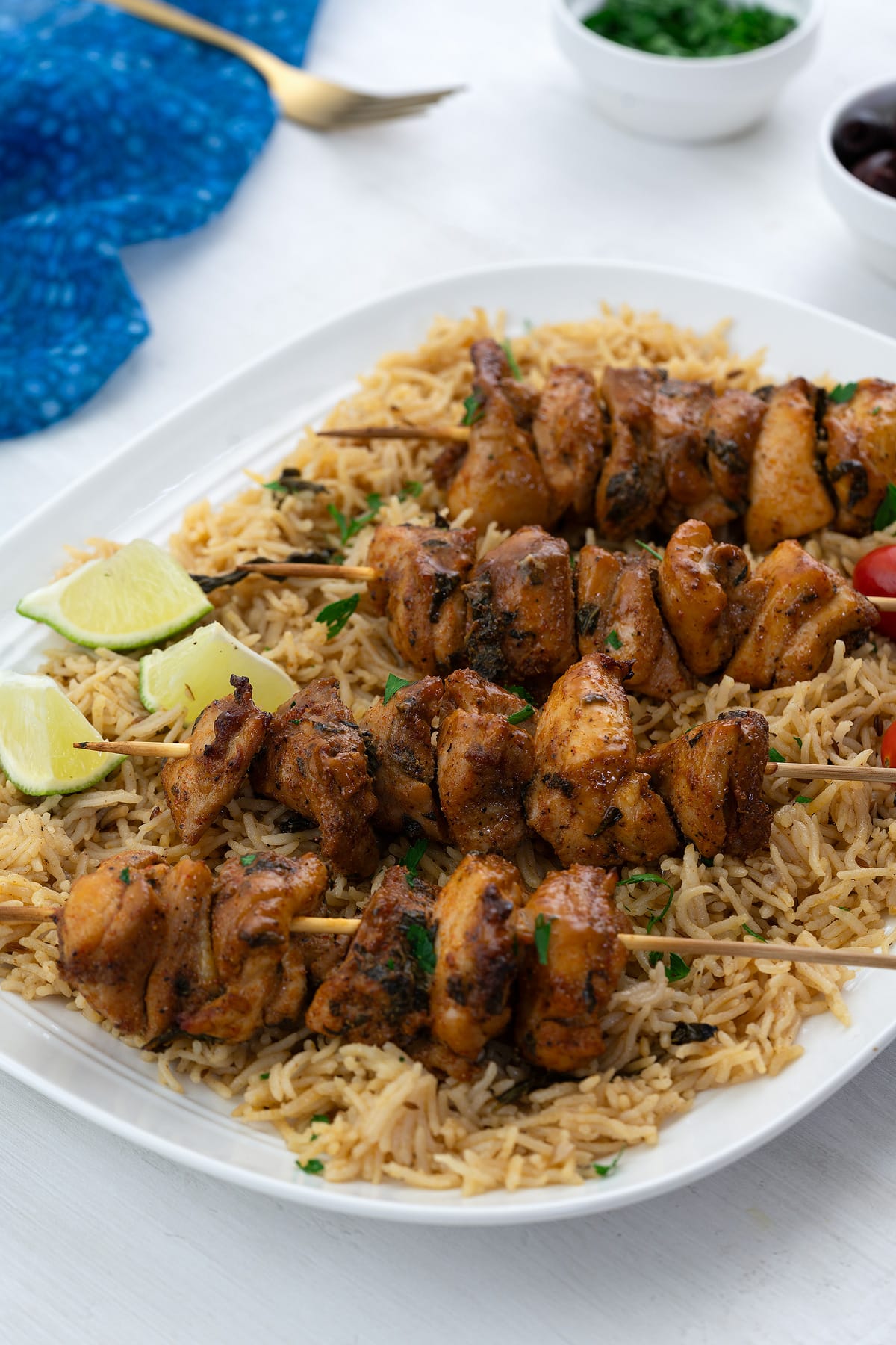 Homemade chicken skewers on a white plate with rice, a golden fork, a blue towel, a small cup of cilantro, and olives on a white table.