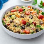 Homemade Mediterranean couscous salad in a white bowl on a white table, surrounded by a blue towel, parsley leaf, grape tomato, and a small cup of black pepper powder.