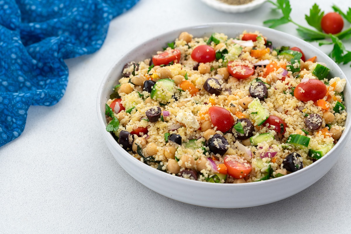 Homemade Mediterranean couscous salad in a white bowl on a white table, surrounded by a blue towel, parsley leaf, grape tomato, and a small cup of black pepper powder.