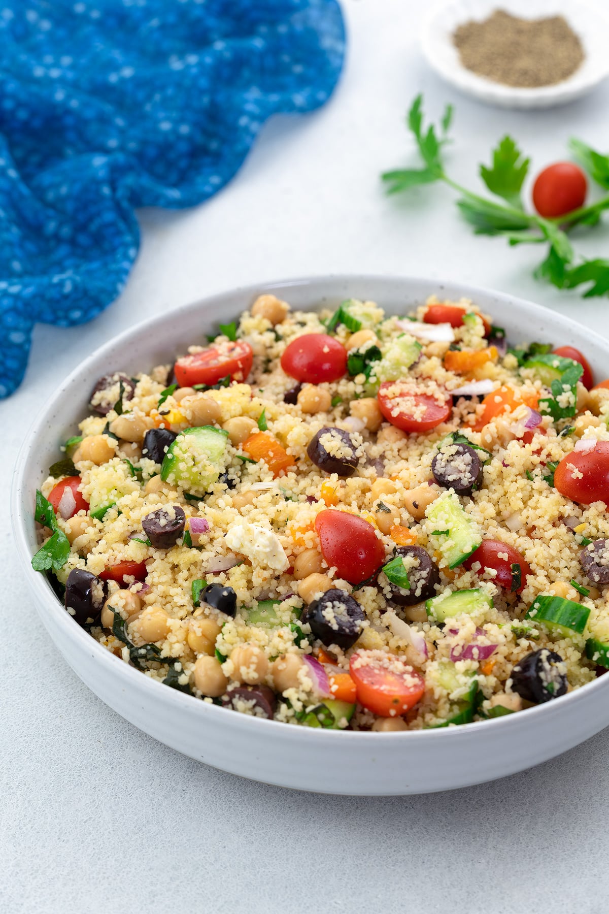 Homemade Mediterranean couscous salad in a white bowl on a white table, surrounded by a blue towel, parsley leaf, grape tomato, and a small cup of black pepper powder.