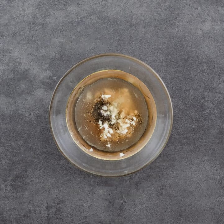 A close-up of a bowl filled with olive oil, red wine vinegar, minced garlic, and spices, ready to be whisked into a vinaigrette dressing.